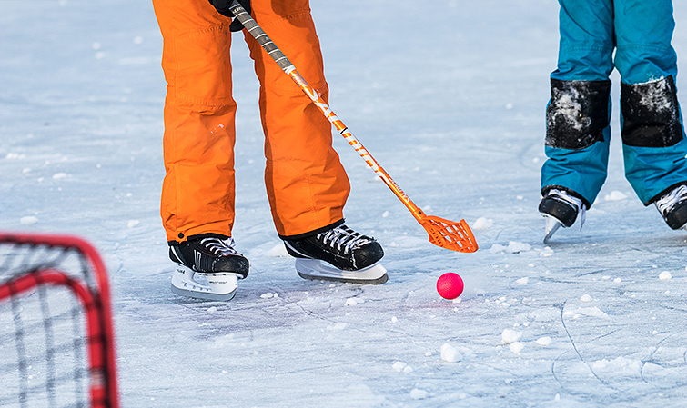 Två skridskoåkare som använder innebandyklubba och bandyboll på isen.